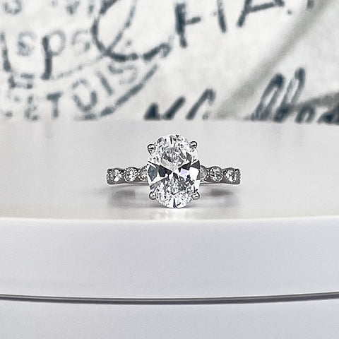 A 4 carat oval diamond engagement ring with pear cut and round diamond accents halfway around the band of 14k white gold and delicate miligran beads framing each small diamond. a vintage inspired ring design.