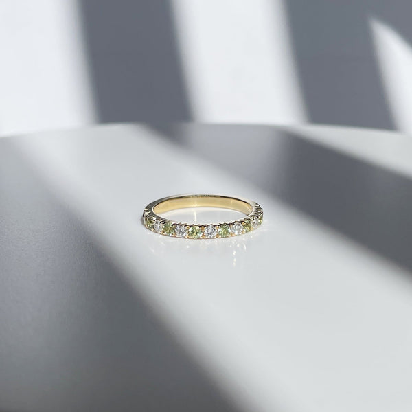 A stunning half eternity ring lays on a white platform with streaking shows framing it and going off into the distance white a beam of light lights up the sapphire and diamond ring. green sapphires, diamonds, pave setting, yellow gold, custom-made, handmade, goldsmith in Vancouver, Gastown jeweller, rings made in Canada 