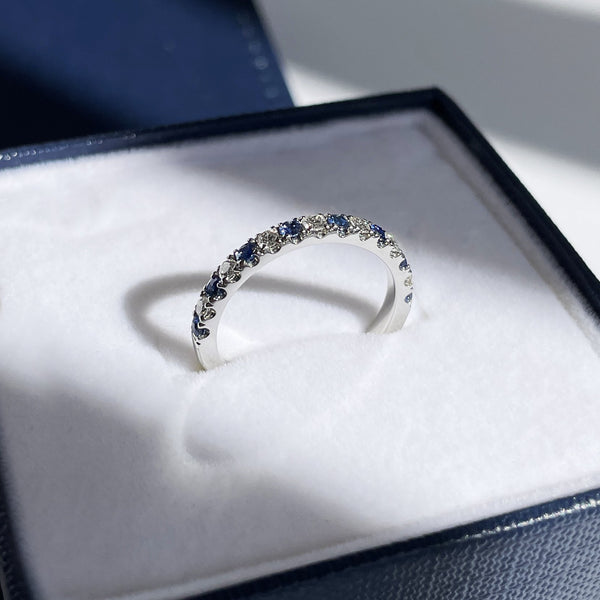 Shadows create depth around a diamond and sapphire eternity band sitting in a blue and white ring box in a beam of light. Pave, eternity band, anniversary band, half eternity ring, u cut pave, lue sapphires, sapphire and diamond ring, natural diamonds, SI1 diamonds, Ceylon sapphires