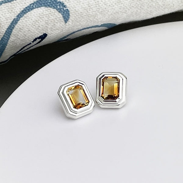 A pair of emerald cut citrine gems set as art deco stud earrings in sterling silver, displayed on a white surface.